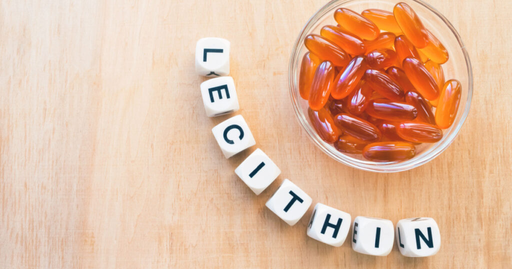 Lecithin gel pills in a round glass bowl and with the word Lecetin from the letters of cubes on a light wooden background. Soy and sunflower lecithin benefits for skin, digestion, lower cholesterol.