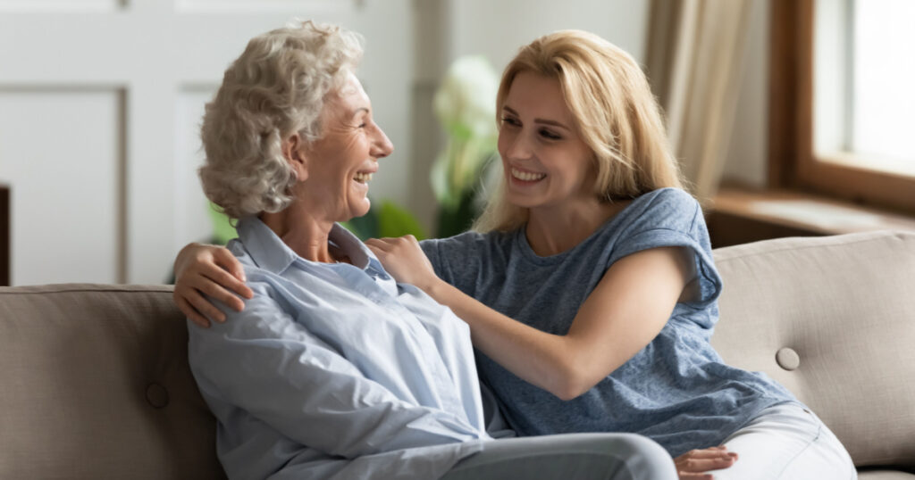 Smiling mature 70s mom and millennial girl child sit on couch in living room laugh talk on weekend together, happy senior mother and adult daughter rest on sofa at home, have fun chatting