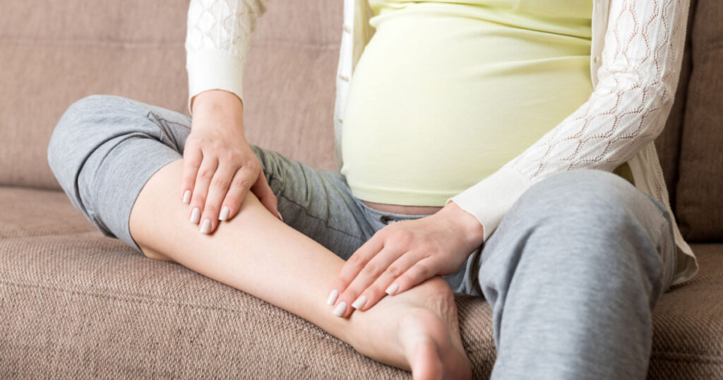 Leg cramps during pregnancy. Closeup of hands massaging swollen foot while sitting on sofa.