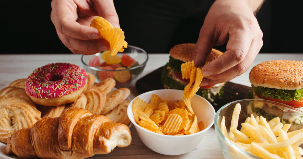 Unhealthy snack, junk food, compulsive overeating. Man overeating unhealthy meals taking hamburger and potato chips from plate
