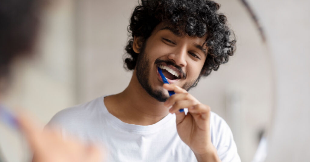 Oral care concept. Young indian man cleaning teeth with toothbrush, smiling to his reflection in mirror, doing toothcare hygiene routine in the morning in bathroom
