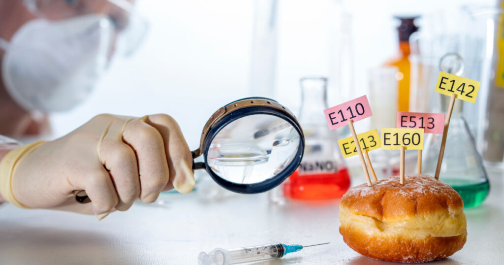In the laboratory assistant's hand is a magnifying glass, on the laboratory table is a donut decorated with tablets with the names of E. Food Laboratory additives.
