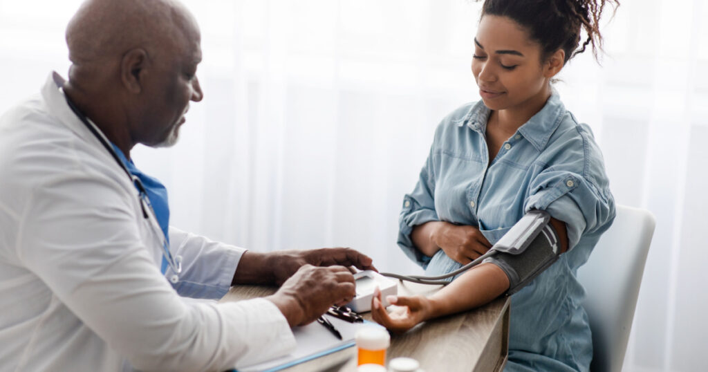 Gestational Hypertension. Mature Male Medical Worker Measuring Arterial Blood Pressure Of Pregnant Black Woman Using Cuff, Patient Having Problems With Tension, Sitting At Table. Health Care Concept
