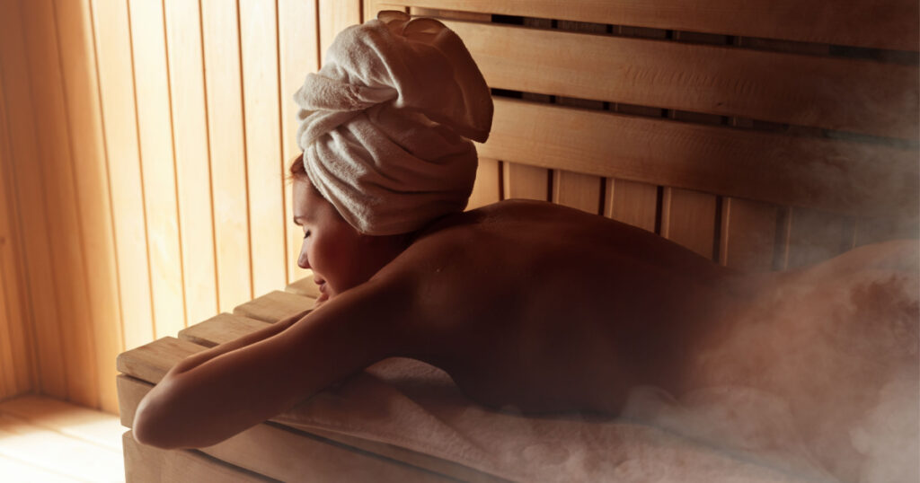 Young woman relaxing and sweating in hot sauna wrapped in towel. Girl In Sauna. Interior of Finnish sauna, classic wooden sauna with hot steam. Russian bathroom. Relax in hot bathhouse with steam.