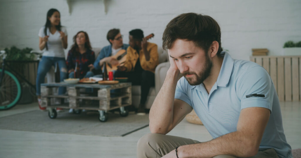 Young student guy feels upset and isolated while his friends celebrating party at home
