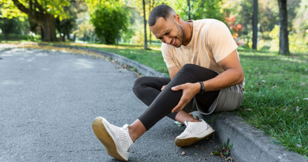 Runner injured his leg while jogging in the park near the trees, hispanic man has muscle spasm massaging his knee, man is running and exercising fitness on a sunny day.