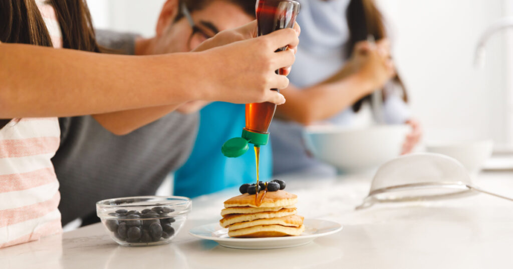 Asian family preparing breakfast in kitchen,father watching daughter put syrup on pancakes. family enjoying preparing meal together at home.
