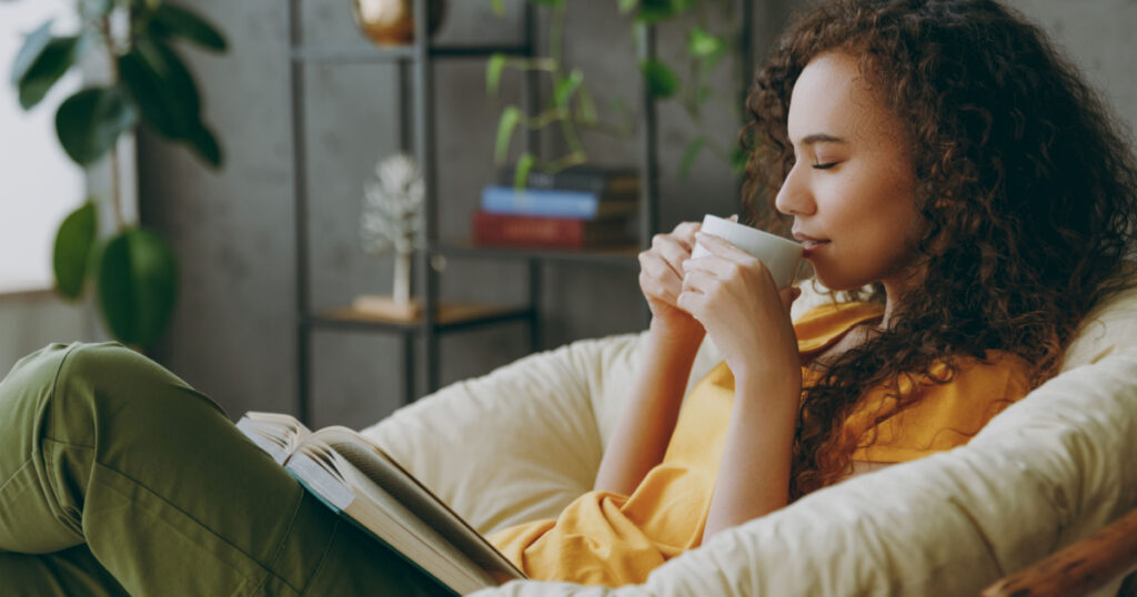 Sideways young woman of African American ethnicity wear t-shirt drink coffee read book close eyes sits in armchair stay at home flat rest relax spend free spare time in living room indoor grey wall