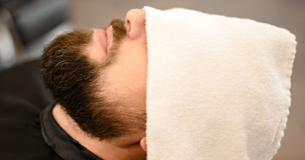 A client is lying on a chair in a barbershop with his beard wrapped in a towel. A towel covers the mans eyes.
