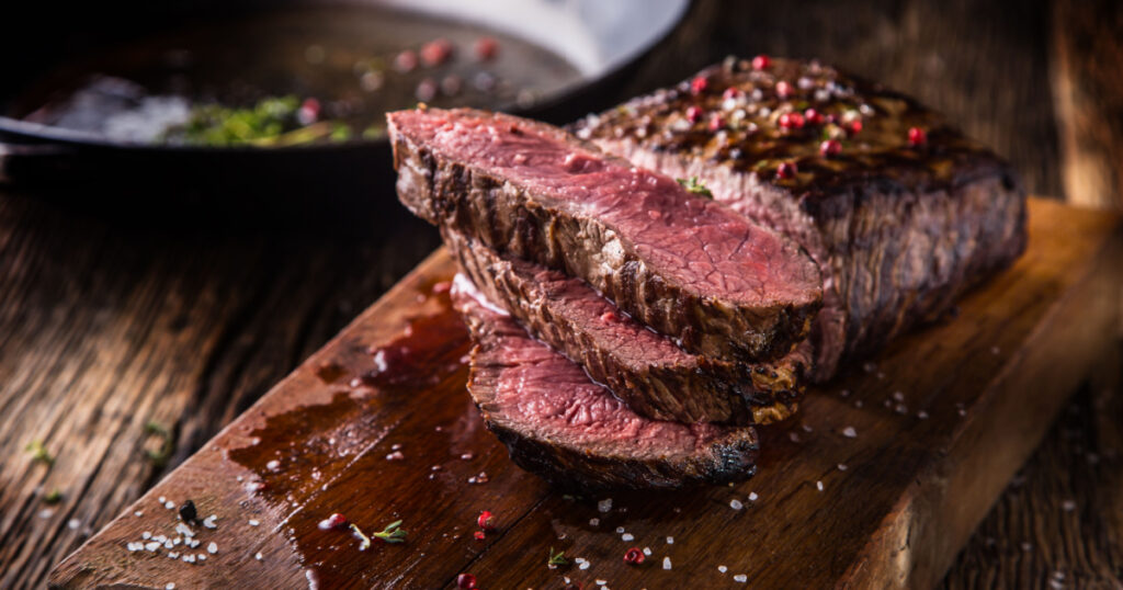 Juicy medium Beef Rib Eye steak slices in pan on wooden board with fork and knife herbs spices and salt.