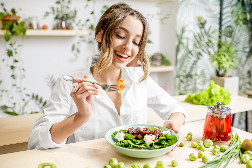 Young woman eating healthy food sitting in the beautiful interior with green flowers on the background
