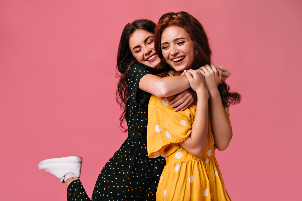 Curly girls smiling and hugging on pink background. Beautiful girlfriends with fashionable make-up in light dresses smile on isolated

