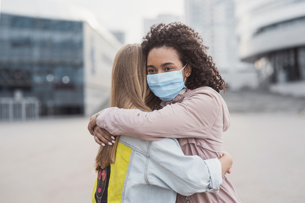 Meeting of two happy friends hugging in the street wearing protective face mask. Multiethnic young women embrace each others in a city during corona virus pandemy
