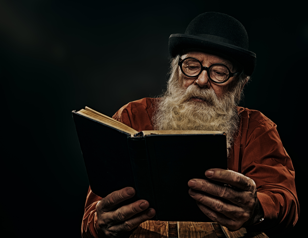 A wise old man with a long gray beard in a bowler hat and glasses is reading an old book. Black background.