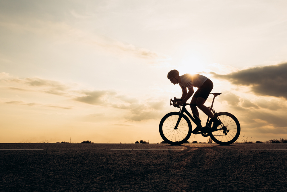 Side view of active sportsman in protective helmet riding bike among nature during sunset. Muscular guy in activewear cycling on paved road.