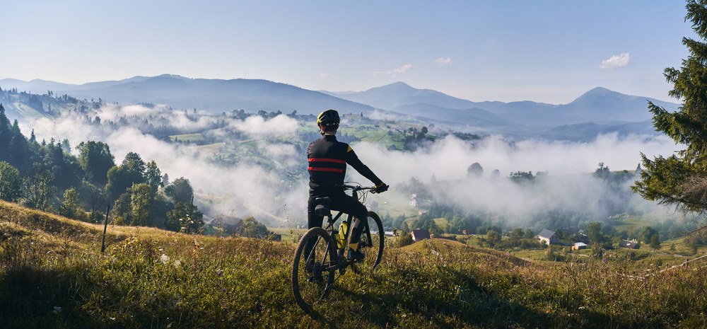 Man riding bicycle on grassy hill and looking at beautiful misty mountains. Male bicyclist enjoying panoramic view of majestic mountains during bicycle ride. Concept of sport, bicycling and nature.