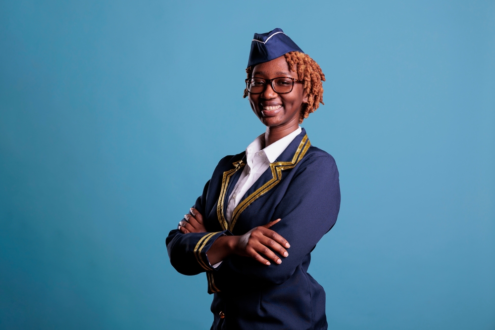 Closeup view of happy african american flight attendant looking with big smile at camera. Flight attendant with optimistic expression smiling in studio shot against blue background.