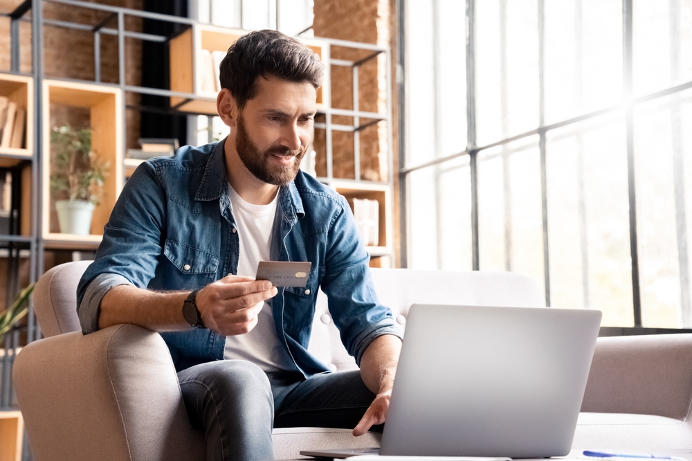 Happy smiling man buying, shopping or paying online with bank card and laptop. Male customer doing internet purchase using web banking service looking at computer screen making secure payment