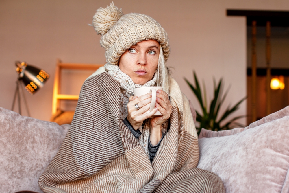 Frozen. Sad woman sit on couch at freezing cooled house in warm cap and blanket shiver tremble with cold. Unhappy middle aged lady spend time at home feel bad suffer of heating system problems
