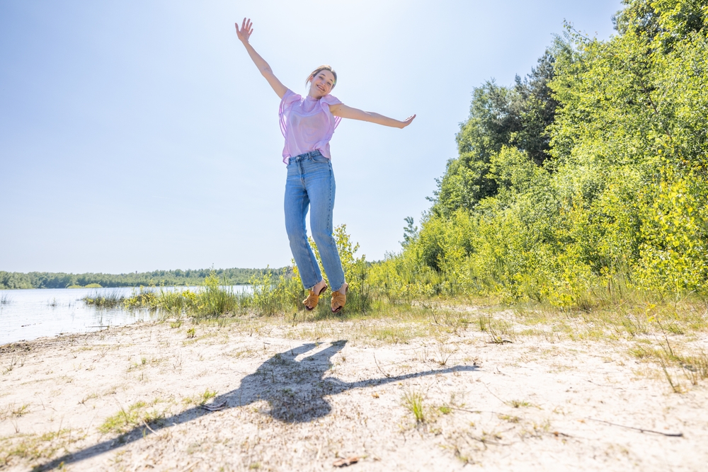 The image captures an exuberant moment as a person joyfully leaps into the air. The individual's shadow is sharply cast on the ground, suggesting the bright and clear conditions of a sunny day. They