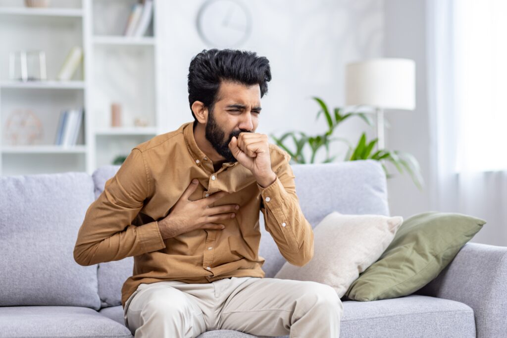 Indian young man sitting at home on the couch and coughing, covering his mouth, holding his chest, feeling severe pain, suffering from an infectious disease.