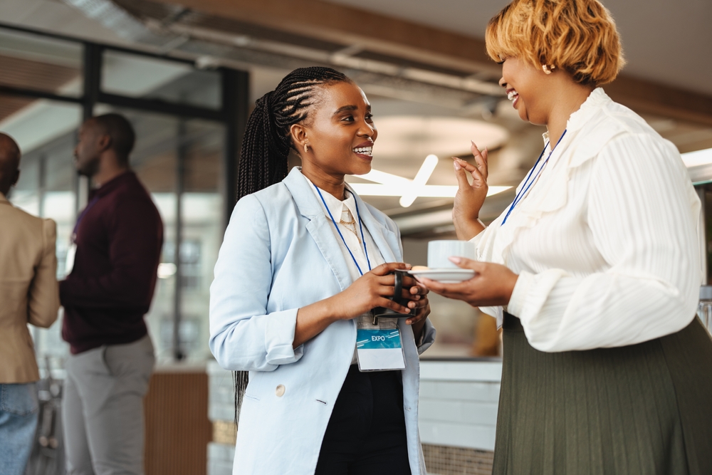 Two professional African businesswomen are engaged in a conversation at a business conference. They hold coffee cups and wear conference badges, smiling as they network in a modern corporate setting.
