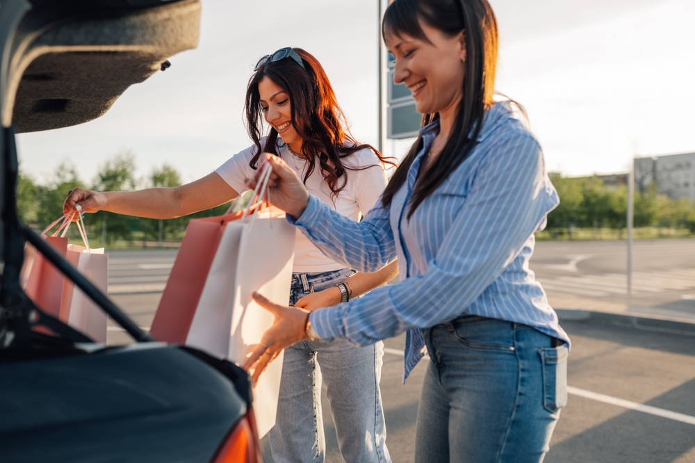 Smiling fashionable female shopper friends standing on parking lot near car trunk and putting paper shopping bags into it. Satisfied trendy chic girls had fun on shopping spree at shopping center.