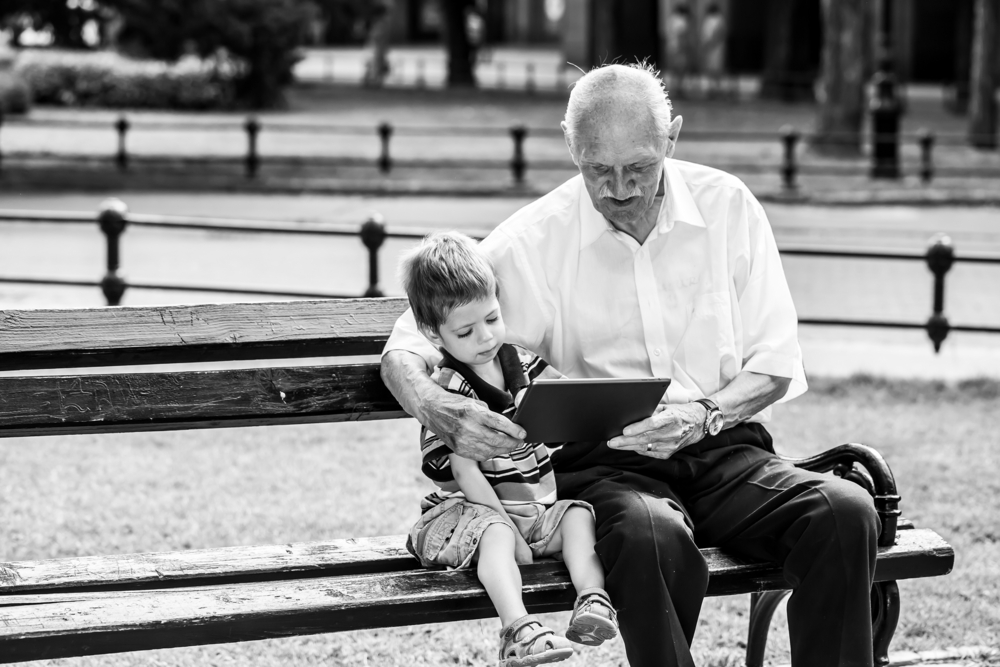 Grandchild teaching to his grandfather to use tablet on a bench. Black and white photography