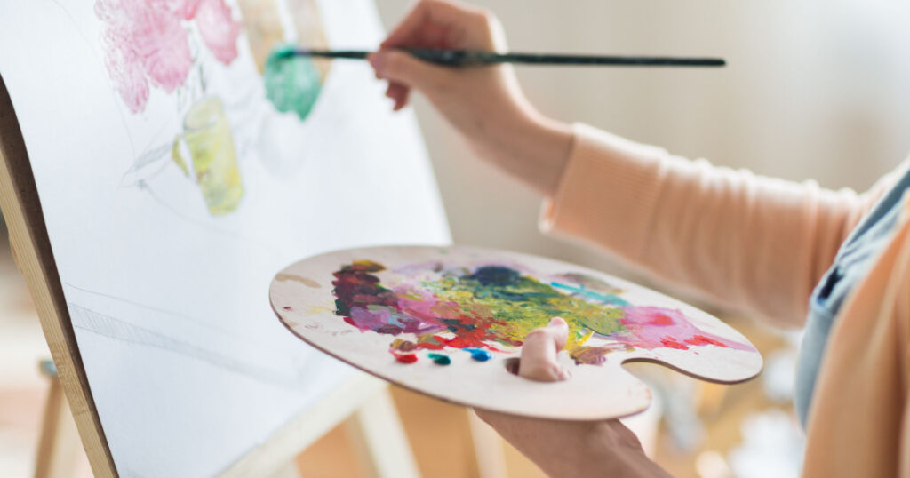 art, creativity and people concept - close up of artist with palette and brush painting still life on paper at studio