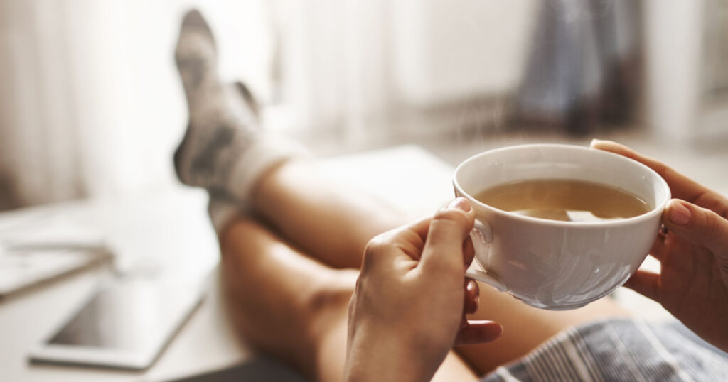 Cup of tea and chill. Woman lying on couch, holding legs on coffee table, drinking hot coffee and enjoying morning, being in dreamy and relaxed mood. Girl in oversized shirt takes break at home