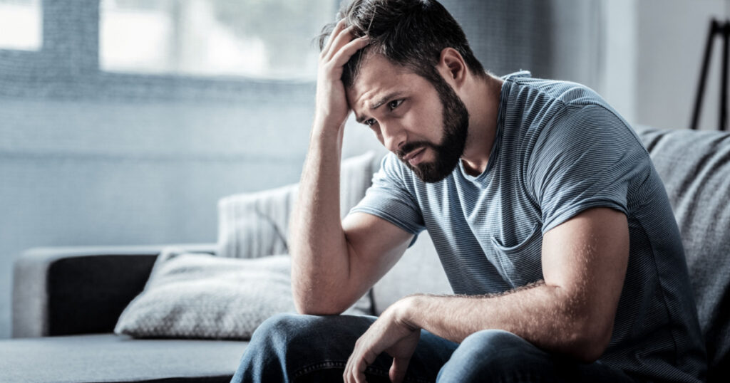 Unpleasant pain. Sad unhappy handsome man sitting on the sofa and holding his forehead while having headache
