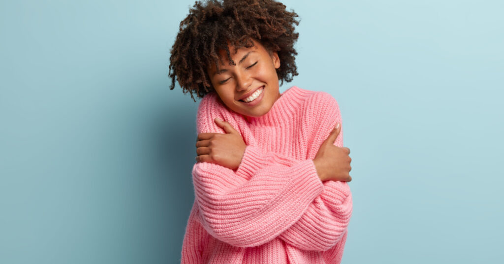 Love yourself concept. Photo of lovely smiling woman embraces herself, has high self esteem, closes eyes from enjoyment, likes her new comfortable soft pink sweater, tilts head, stands indoor