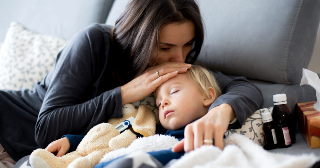 Blond toddler boy, sleeping on the couch in living room, lying down with fever, mom checking on him