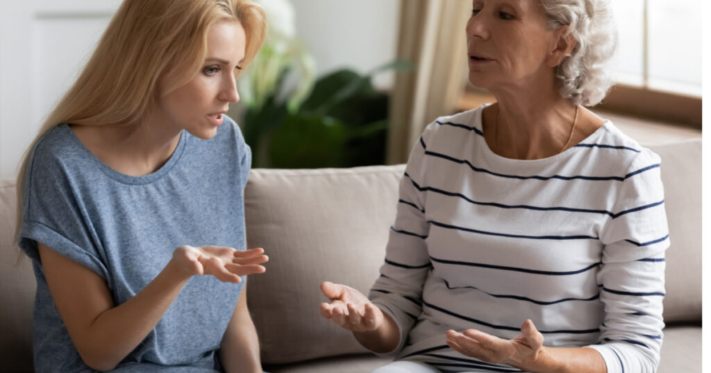 Stressed young blonde grown up daughter arguing with nervous old mature mother, sitting together at home. Irritated elderly woman lecturing adult child, different generations misunderstanding gap.