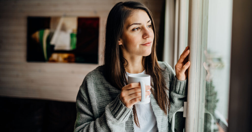 Young woman spending free time home.Self care,staying home.Enjoying view,gazing through to the window.Quarantined person indoors.Serene mornings.Avoiding social contact.