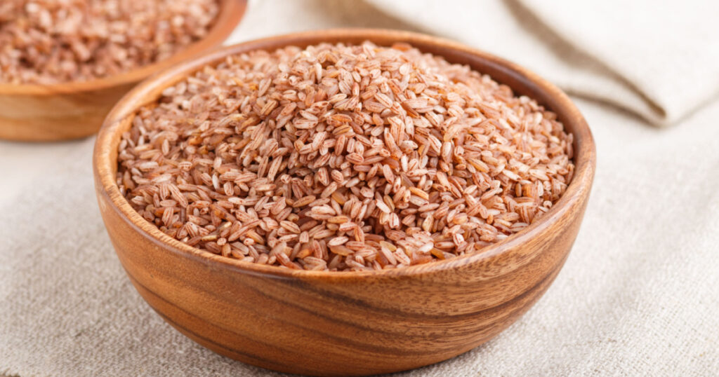 Two wooden bowls with unpolished brown rice on a white wooden background and linen textile. Side view, close up, selective focus.