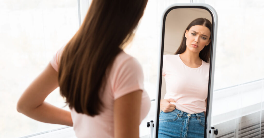 Weight Gain. Unhappy Girl Touching Drooping Belly Standing In Front Of Mirror At Home. Empty Space, Selective Focus