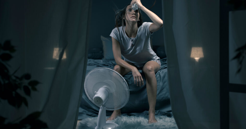 Exhausted woman suffering suring the heatwave, she is holding a water bottle and sitting in front of a cooling fan in the bedroom