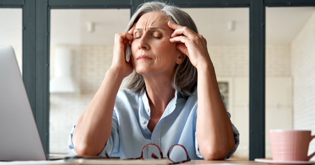 Tired stressed old mature business woman suffering from headache at work. Upset sick senior middle aged lady massaging head feeling migraine from overwork or menopause using computer at home office.
