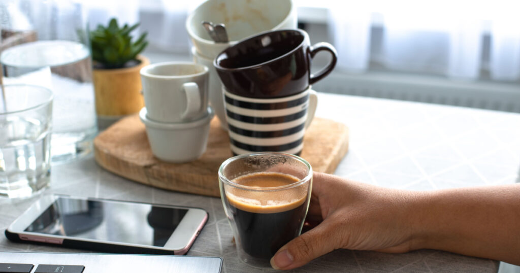 A young female freelancer working from home holding a cup of coffee with many cups in the background drinks too much coffee caffeine addiction anxious and crazy in maniac bad lifestyle concept
