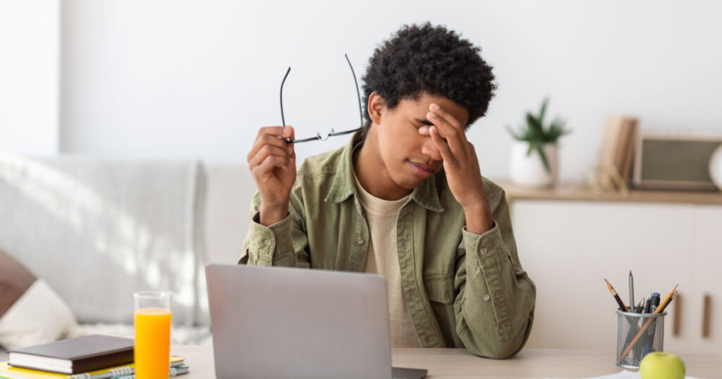 Black teenager feeling tired, rubbing irritated eyes, sitting at desk with laptop, exhausted from studying online. African American guy feeling fatigue from looking at screen, doing assignments on web
