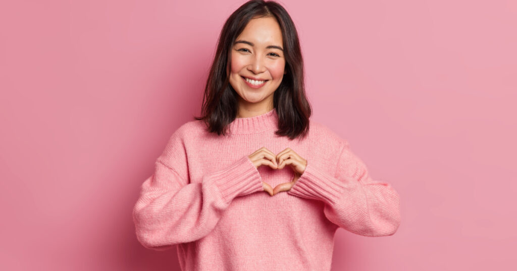 Attractive brunette young Asian woman feels happy and romantic shapes heart gesture expresses tender feelings wears casual jumper poses against pink background. People affection and care concept