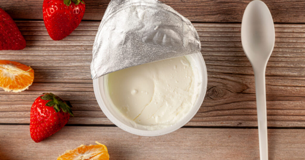Flat lay image of a store bought plastic cup of plain low fat Greek Yogurt with the aluminum foil seal peeled off. It is on wooden background with a plastic spoon and fresh fruits. A diet idea concept