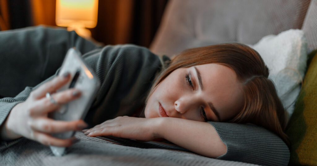 Sad teenage girl suffering from insomnia surfing Internet or chatting using smartphone at night lying on bed. Depression Internet addiction and depression in young woman. Long web banner.