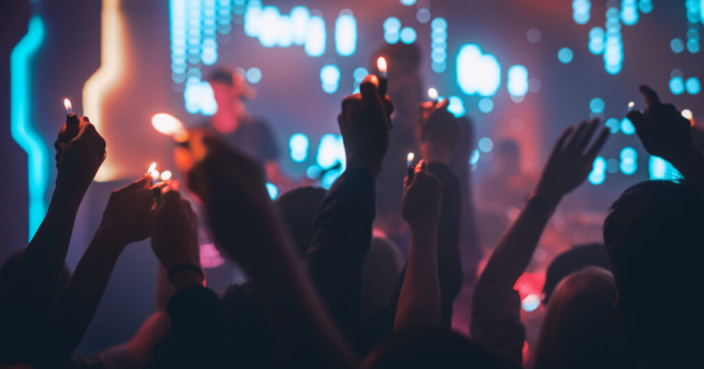 Rock Band Performing a Slow Song at a Concert in a Night Club. Front Row Crowd is Holding Lighters. Silhouettes of Fans Raise Hands in Front of Bright Colorful Strobing Lights on Stage.