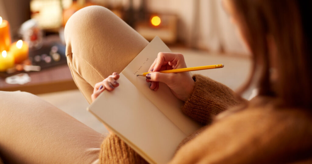 halloween, holidays and leisure concept - close up of young woman with pencil writing to diary and resting her feet on table at cozy home