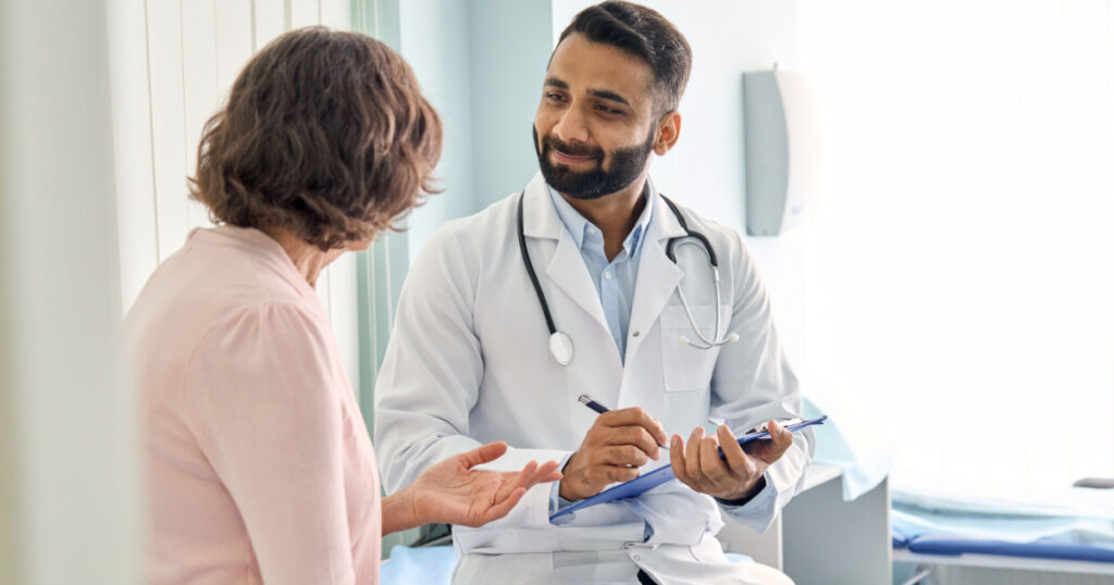 Indian male doctor consulting senior old patient filling form at consultation. Professional physician wearing white coat talking to mature woman signing medical paper at appointment visit in clinic.