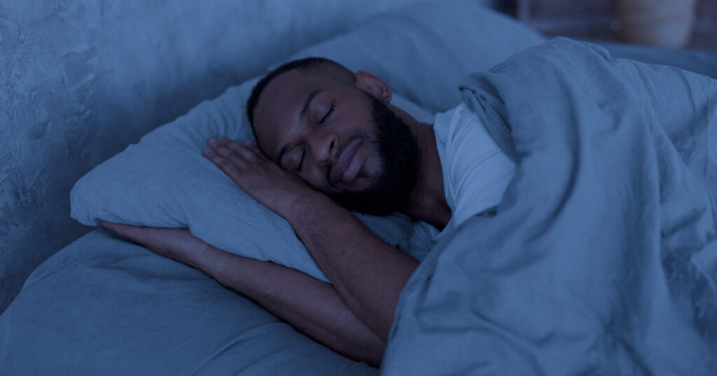 Healthy Sleeping Concept. Portrait of happy young well-slept African American man lying in bed with closed eyes, resting in bedroom on the side in the dark night, having hap