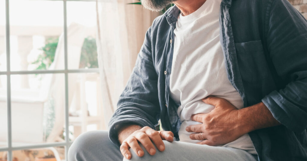 Man suffering with severe stomach pain sitting at home. Hand of mature guy holding abdomen suffering from ache, diarrhea or indigestive problem. Caucasian guy pressing on belly on painful sensation