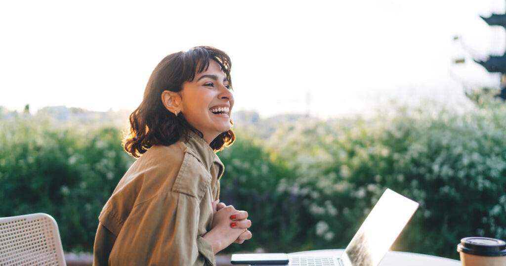 Side view of young brunette smiling happily while sitting at table with laptop working remotely on new web design project in sunny garden